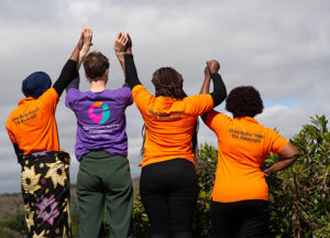 Mehrere Personen stehen mit dem Rücken zur Kamera und strecken gemeinsam die Arme in die Höhe; auf den Shirts sind „Opportunity International Deutschland“ und „Usiwa Watha Project TIA Machinjiri“ zu sehen.